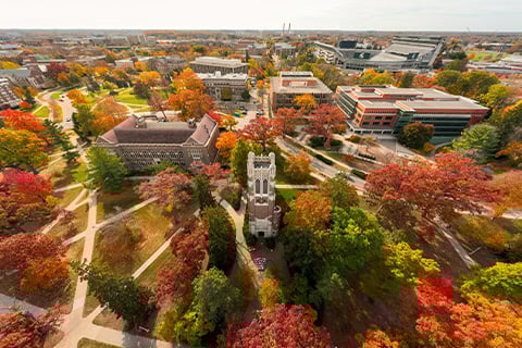 Aerial image of MSU's Beaumont tower in the fall. The changing leaves on the trees are visible along with MSU's network of sidewalks and buildings.