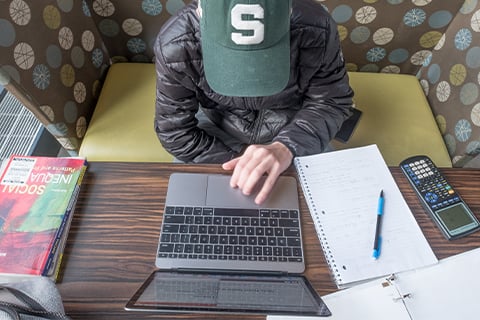 View of a person from above. They are wearing a green ballcap with a block Spartan S in white. They have a long sleeved black jacked on and are working on an open laptop. The laptop keyboard, their notebook, calculator and other school materials are in bold feature.