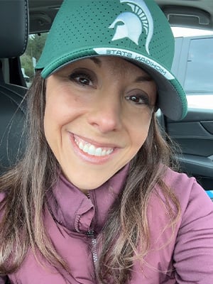 Portrait. The individual is looking directly at the camera and smiling. They are wearing a green MSU ballcap, with the Spartan helmet on the front. Their hair is long brownish-blonde, and hangs over their shoulders. 
