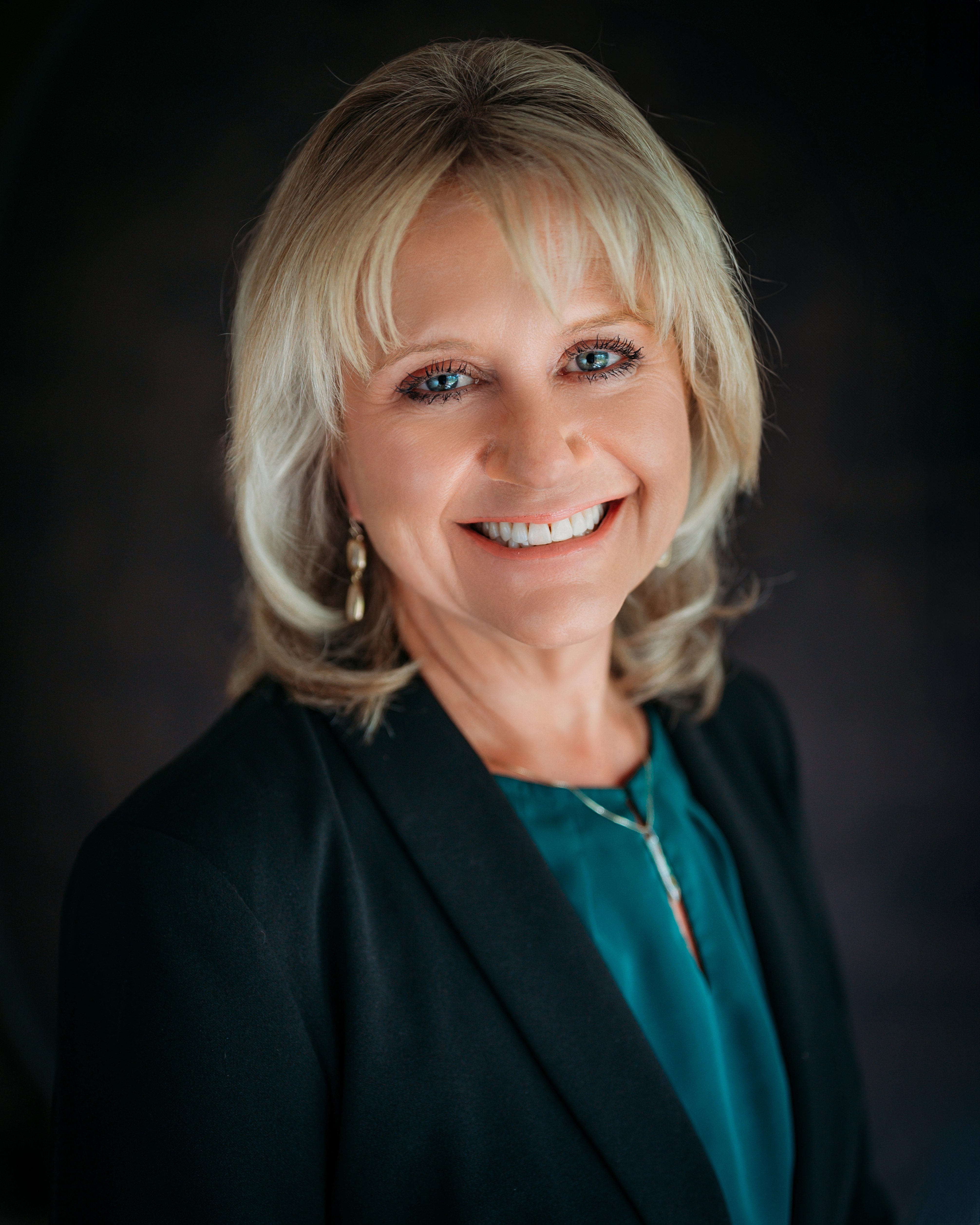 Studio portrait of Amy Martin wearing a dark blazer over a green top. She has short blond hair. 