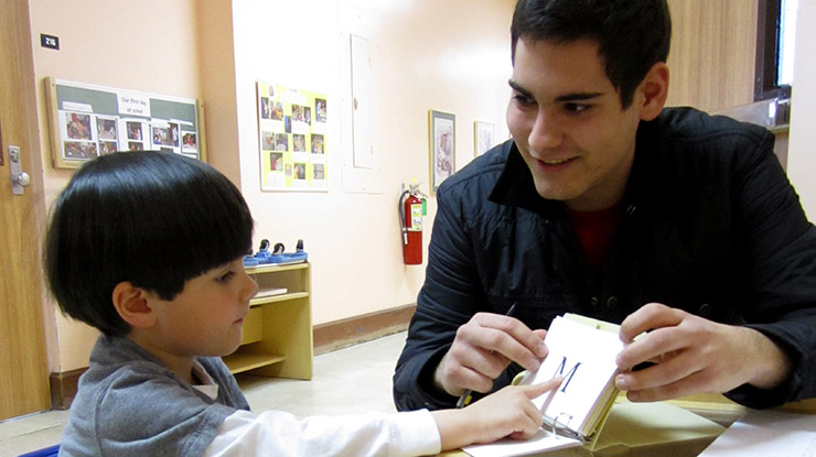 A student pursuing a child development degree sits with a young child who points to the letter ?M? on a teaching card.