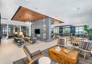 Modern upscale spacious community clubhouse with multiple seating areas, entertainment lounge area with wall-mounted TV and wood beam ceiling with chandelier next to picture windows at MAA Breakwater luxury apartments in Tampa, FL