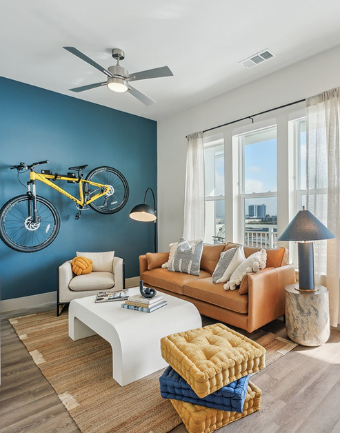 Upscale modern living room with a bicycle hanging on a rack on a blue accent wall, a ceiling fan, orange couch with a white coffee table, a glass door entrance and windows with a waterfront view at MAA Breakwater luxury apartments in Tampa, FL