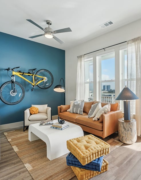 Upscale modern living room with a bicycle hanging on a rack on a blue accent wall, a ceiling fan, orange couch with a white coffee table, a glass door entrance and windows with a waterfront view at MAA Breakwater luxury apartments in Tampa, FL