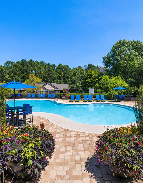 Swimming pool with blue lounge chairs and patio table with sun umbrella surrounded by greenery at MAA Berkeley Lake luxury apartments in Duluth, GA