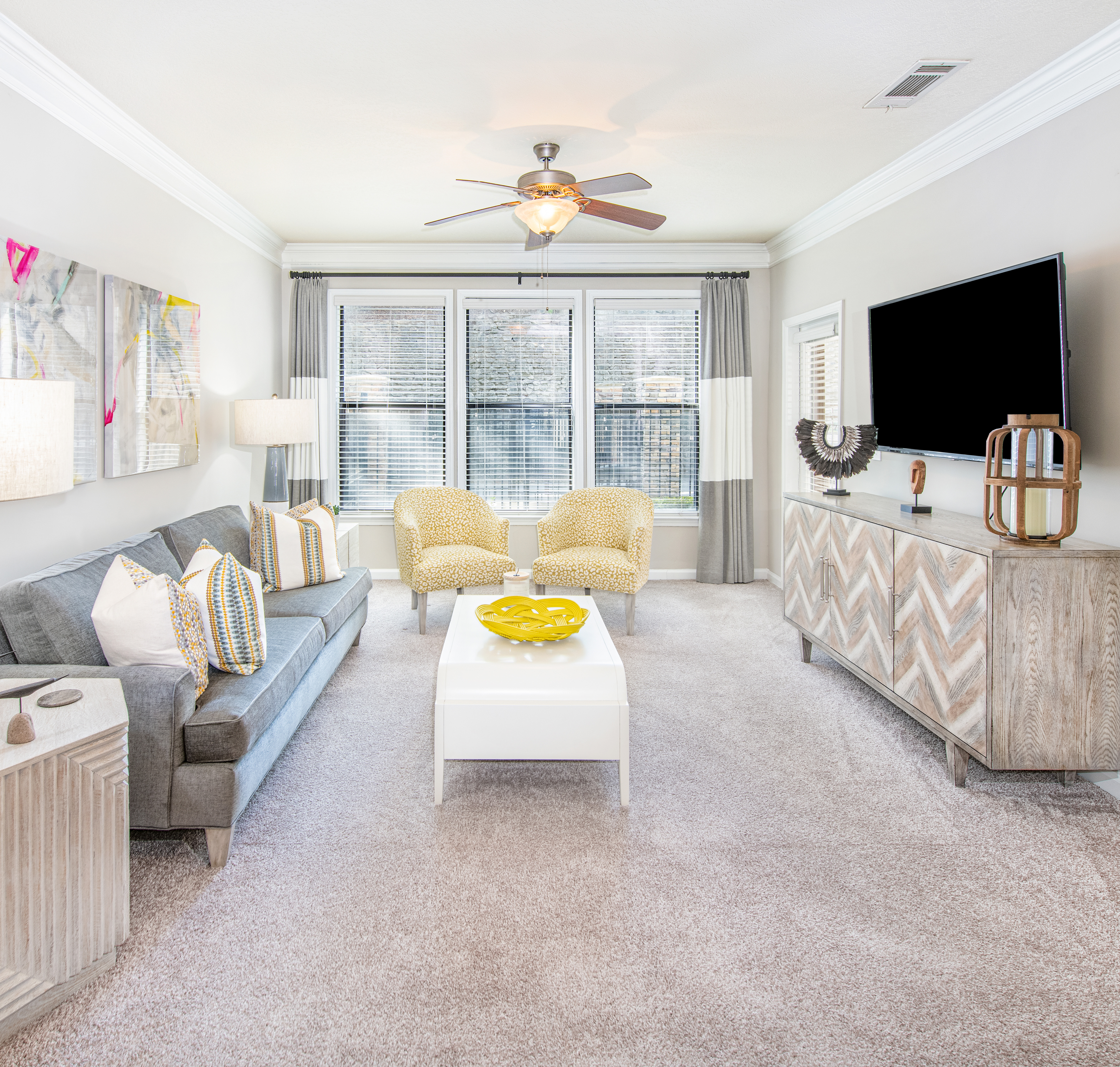 Model apartment living room with a ceiling fan, plush couch sitting area with a white rectangular coffee table and wall-mounted TV next to large picture windows with curtains and blinds at MAA Brookwood luxury apartments in Atlanta, GA