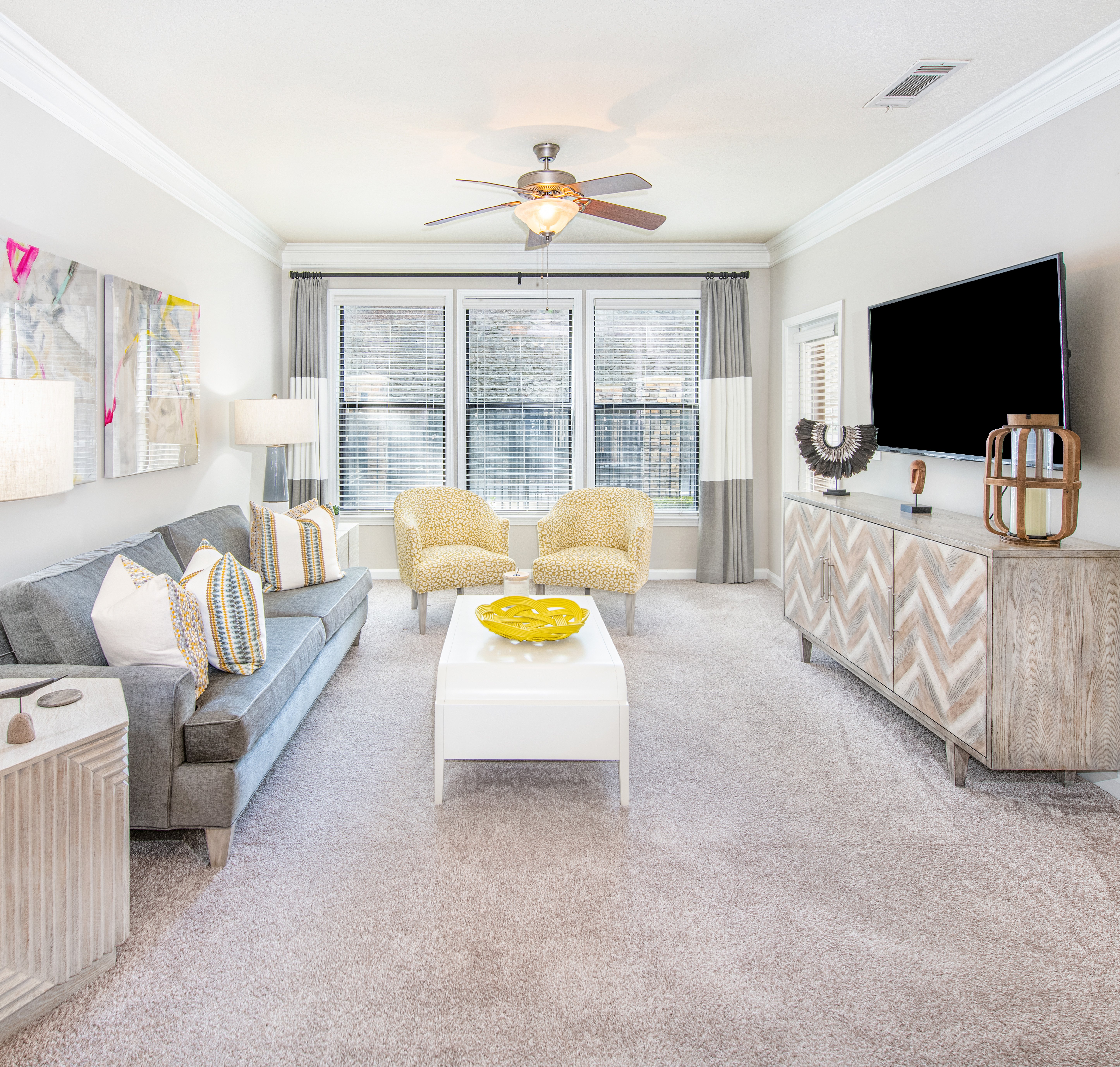 Model apartment living room with a ceiling fan, plush couch sitting area with a white rectangular coffee table and wall-mounted TV next to large picture windows with curtains and blinds at MAA Brookwood luxury apartments in Atlanta, GA