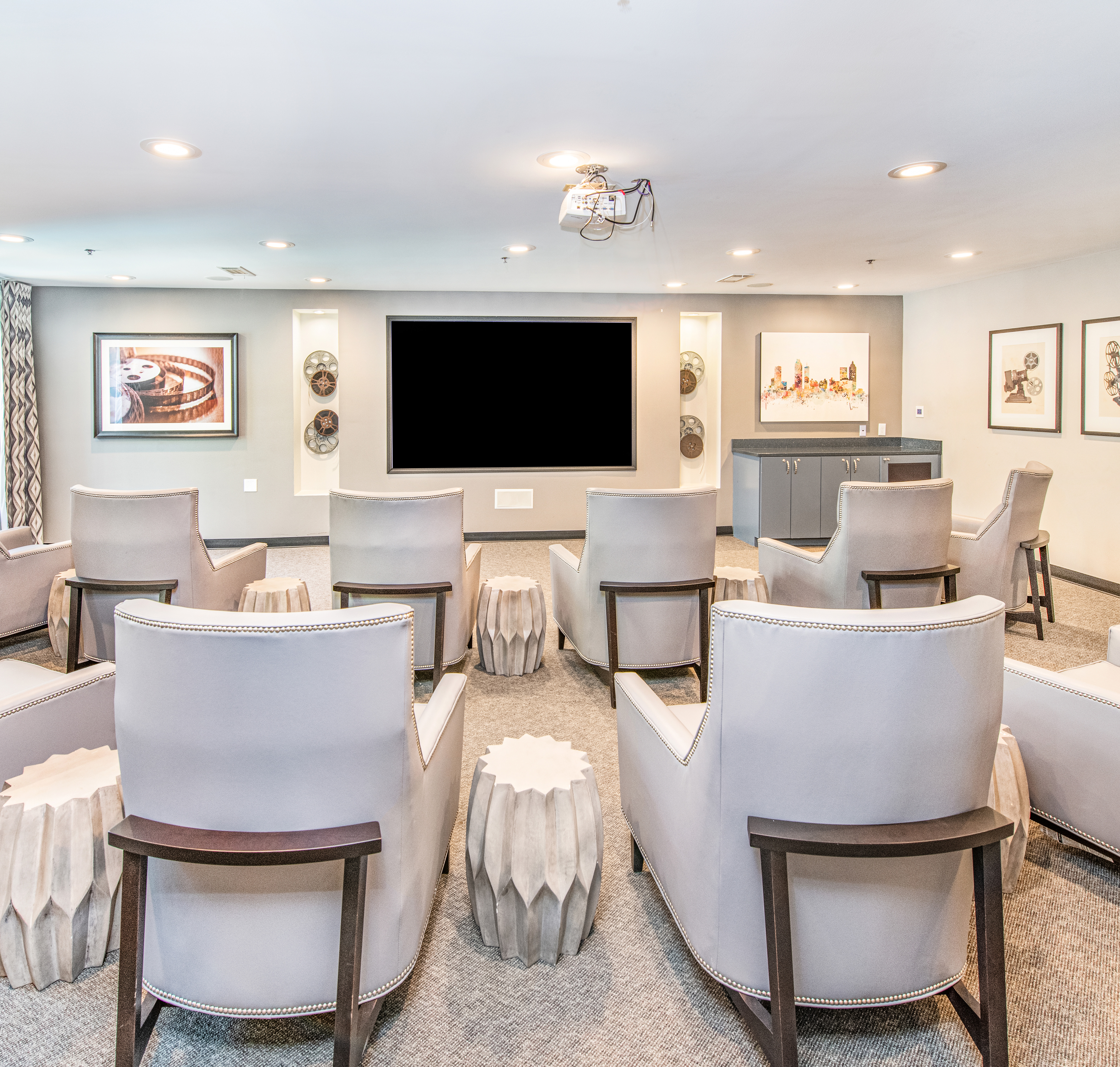 Modern glam-style community private theater with rows of plush white arm chairs and cylinder-shaped tables, a large movie screen and wall art at MAA Brookwood luxury apartments in Atlanta, GA
