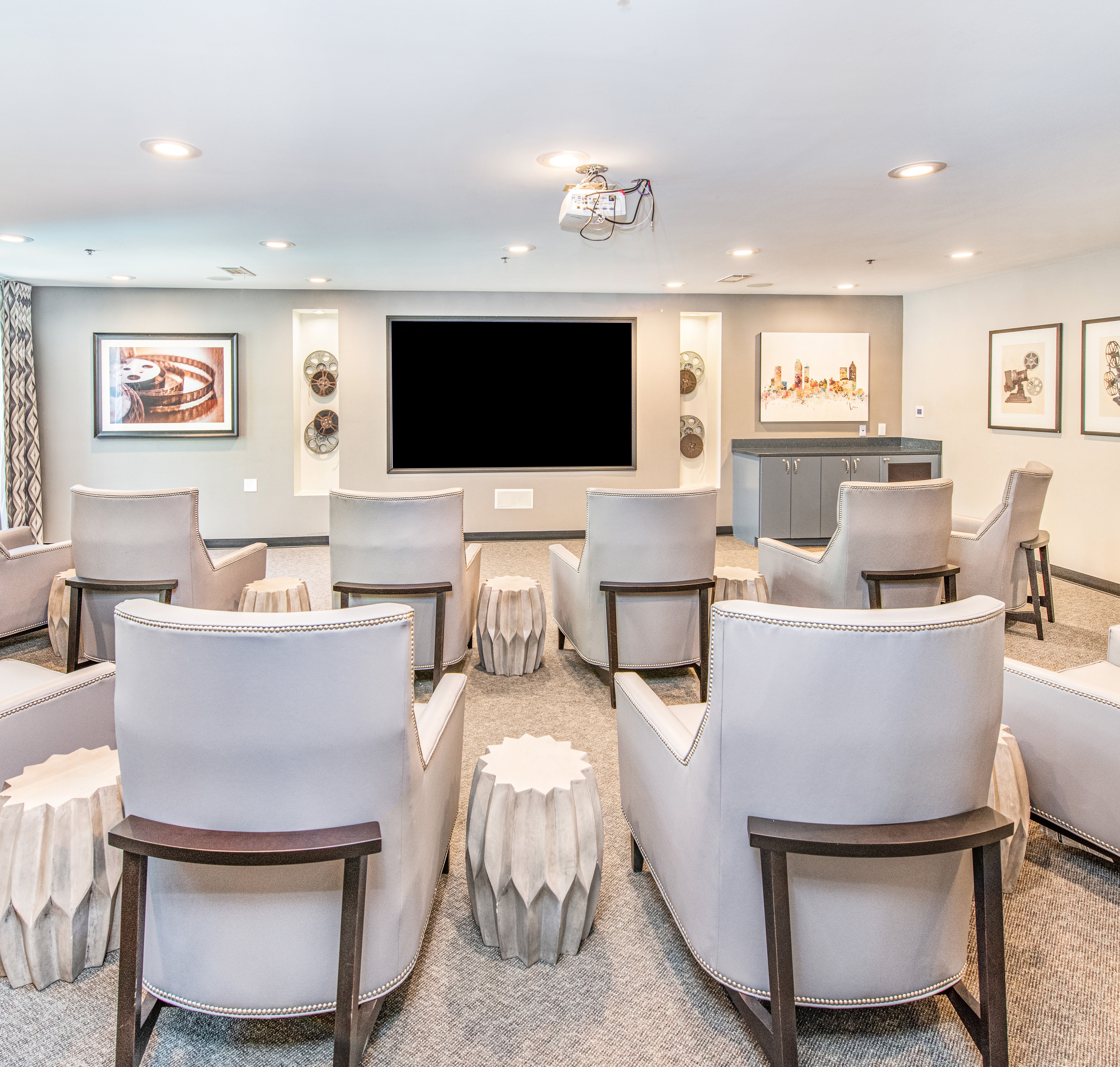 Modern glam-style community private theater with rows of plush white arm chairs and cylinder-shaped tables, a large movie screen and wall art at MAA Brookwood luxury apartments in Atlanta, GA