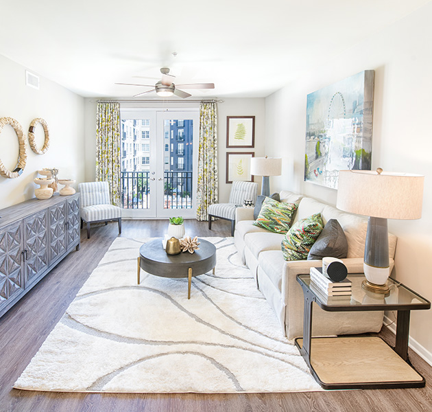 Apartment living room with ceiling fan and double glass doors leading to a balcony view, and a white couch with a circular coffee table on white area rug on wood floors at MAA Buckhead luxury apartments complex in Atlanta, GA
