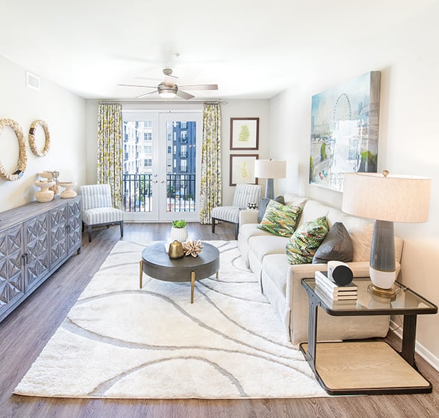 Apartment living room with ceiling fan and double glass doors leading to a balcony view, and a white couch with a circular coffee table on white area rug on wood floors at MAA Buckhead luxury apartments complex in Atlanta, GA