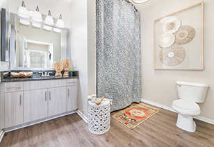 Wide view of modern bathroom with single sink vanity with lights and light wood cabinets, shower curtains and wood floors at MAA Buckhead luxury apartments complex in Atlanta, GA