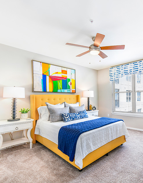 Colorful model bedroom with abstract wall art above bed with headboard and accent pillows, a ceiling fan and windows with carpeted floors at MAA Buckhead luxury apartments complex in Atlanta, GA