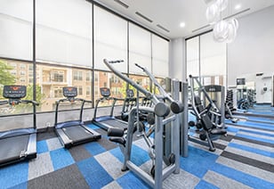 Fitness center with rows of weight machines and treadmills alongside wall of large picture windows with shades on blue carpeted floors at MAA Buckhead luxury apartments complex in Atlanta, GA