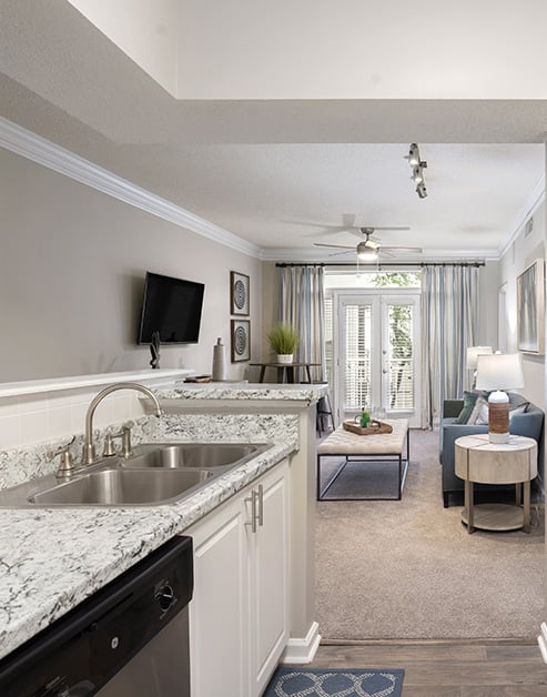 Side view of open kitchen with granite countertops, stainless steel double sink, dishwasher and white cabinets on wood floors next to living area at MAA Gardens luxury apartments in Atlanta, GA