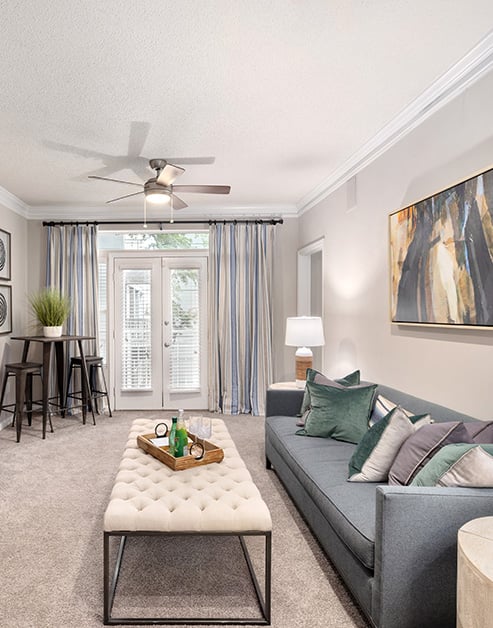 Model modern living room with modern d�cor, ceiling fan next to double glass doors with curtains, a grey sofa couch with accent pillows and wall art with a white ottoman at MAA Gardens luxury apartments in Atlanta, GA