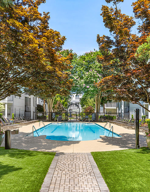 Gated swimming pool with lounge chairs with an arch entrance and a paved walkway with grass surrounded by trees at MAA Gardens luxury apartments in Atlanta, GA