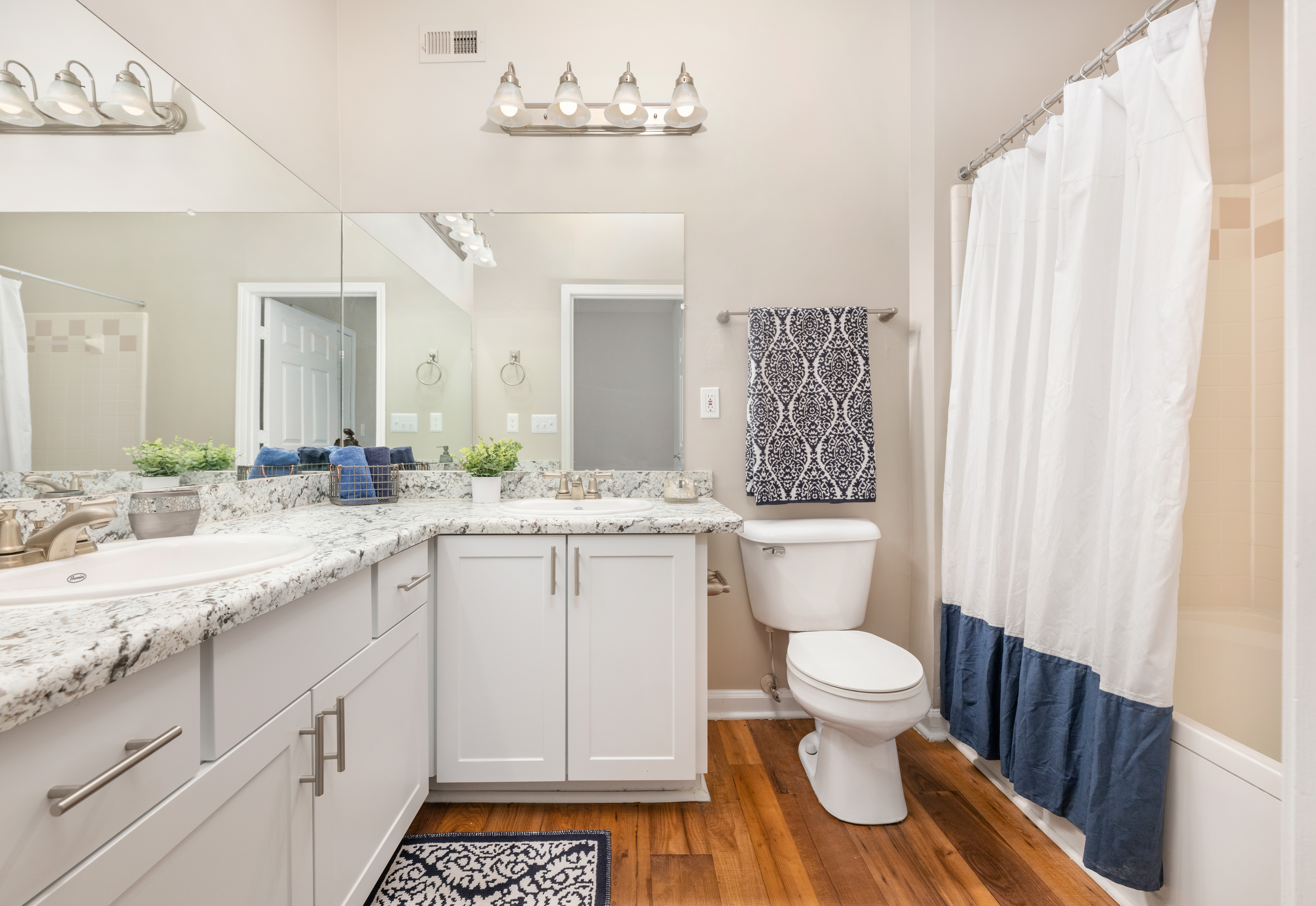 Long view of bathroom with a large vanity mirror and double sinks on light-colored countertops, white cabinets next to a toilet and white blue shower curtains on wood floors at MAA Lake Lanier luxury apartments in Gainesville, GA