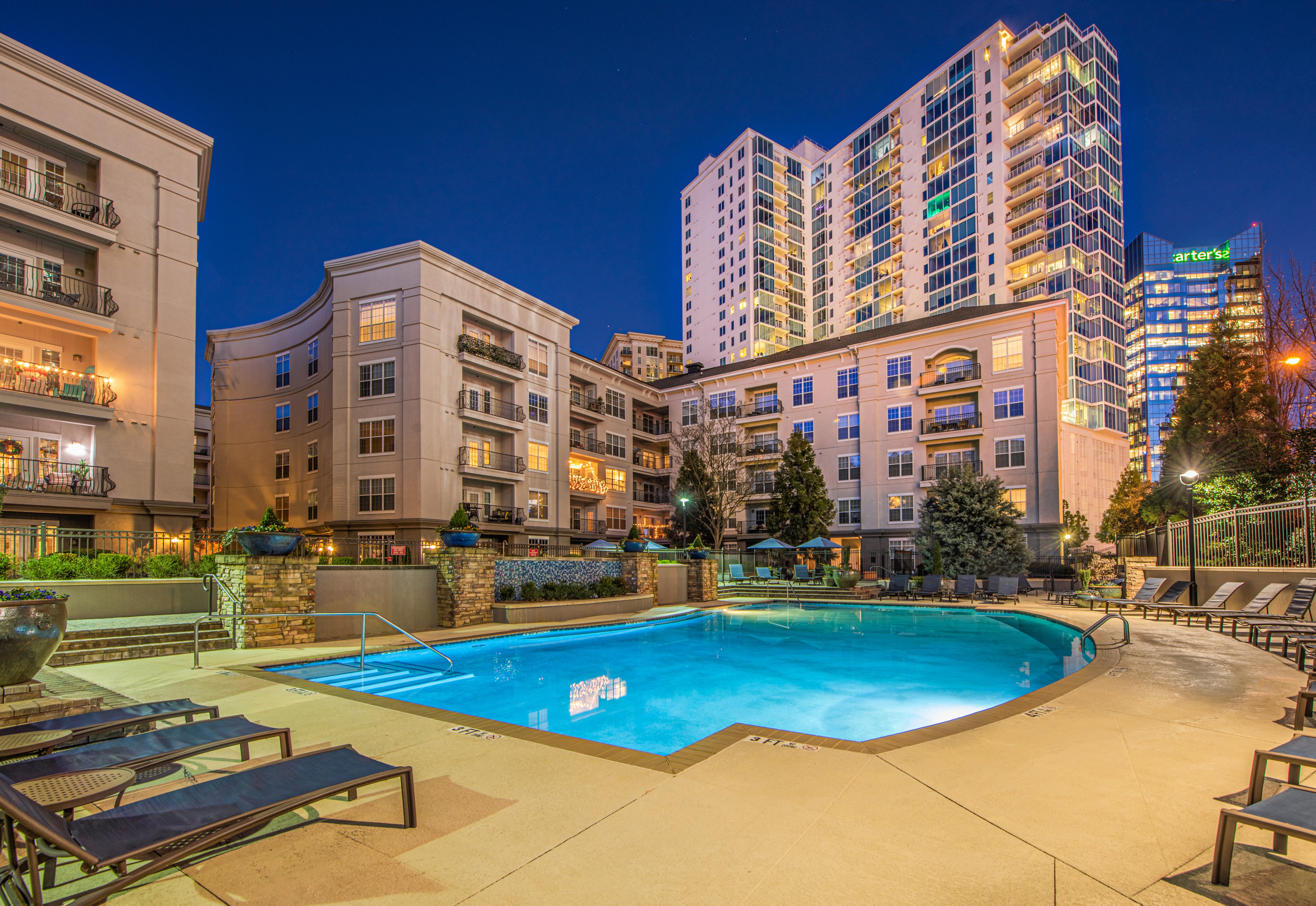 Wide view of a lighted swimming pool at night surrounded by lounge chairs and trees next to buildings with a city skyline view at MAA Lenox luxury apartment homes in Atlanta, GA