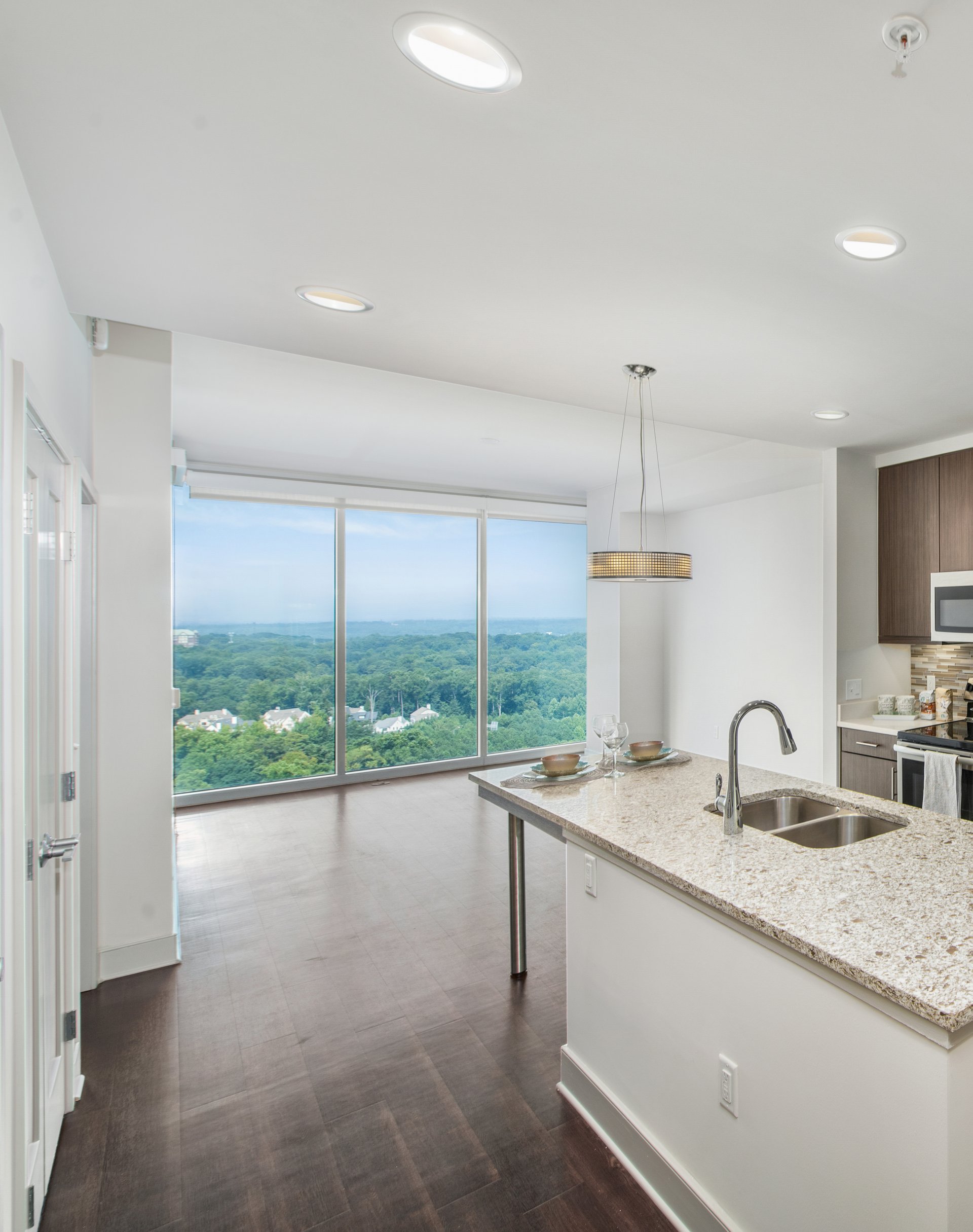 Open living room space with large wall-length picture windows with a skyline view on wood floors next to a kitchen at MAA Lenox luxury apartment homes in Atlanta, GA