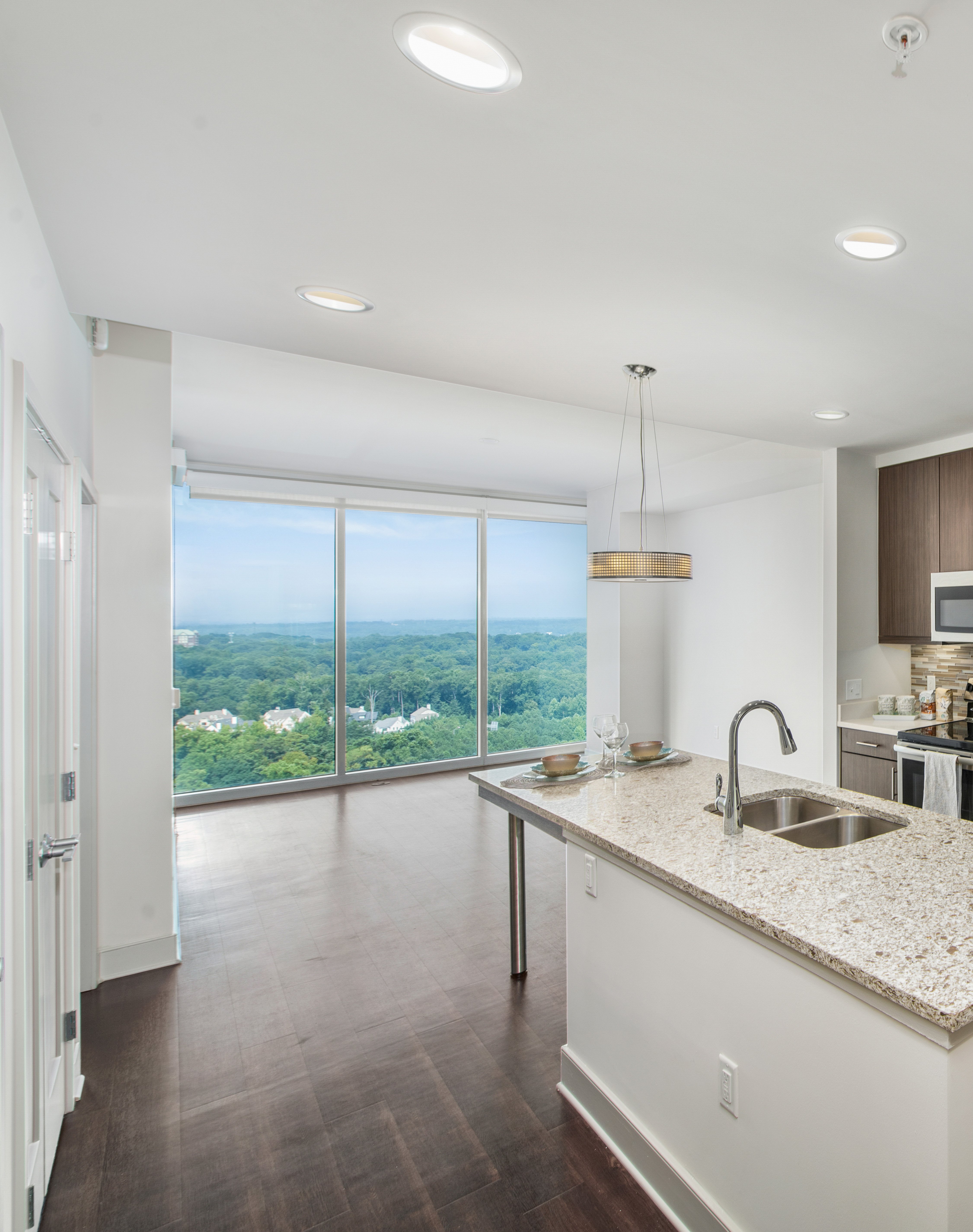 Open living room space with large wall-length picture windows with a skyline view on wood floors next to a kitchen at MAA Lenox luxury apartment homes in Atlanta, GA