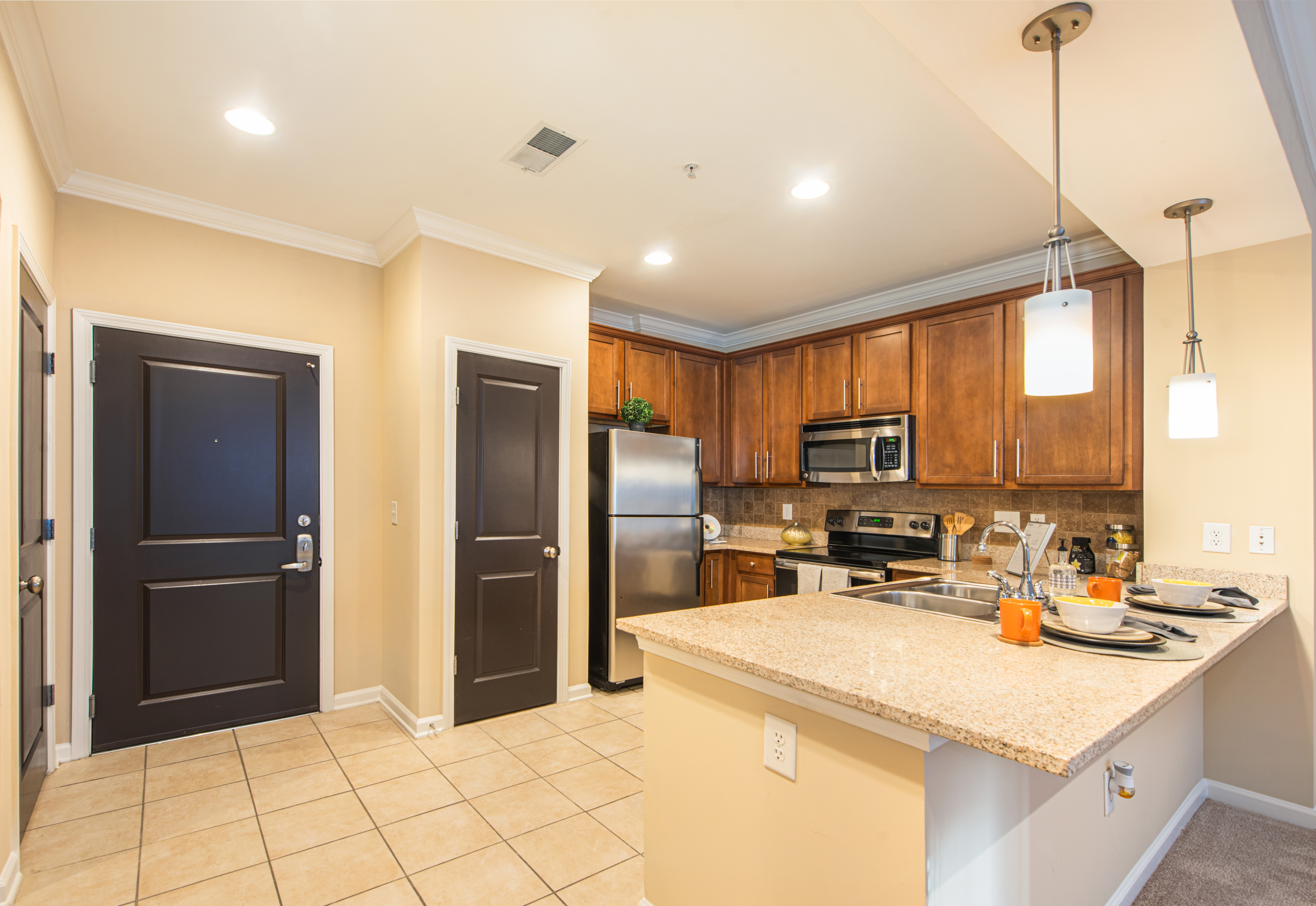 Open kitchen with wood cabinets, hanging light fixtures over a bar top counter with stainless steel appliances on tiled floors at MAA Lenox luxury apartment homes in Atlanta, GA