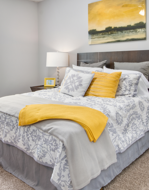 Cozy model bedroom with grey toned bedding, yellow accent pillow and throw blanket, and wall art at MAA Milstead luxury apartments in Kennesaw, GA