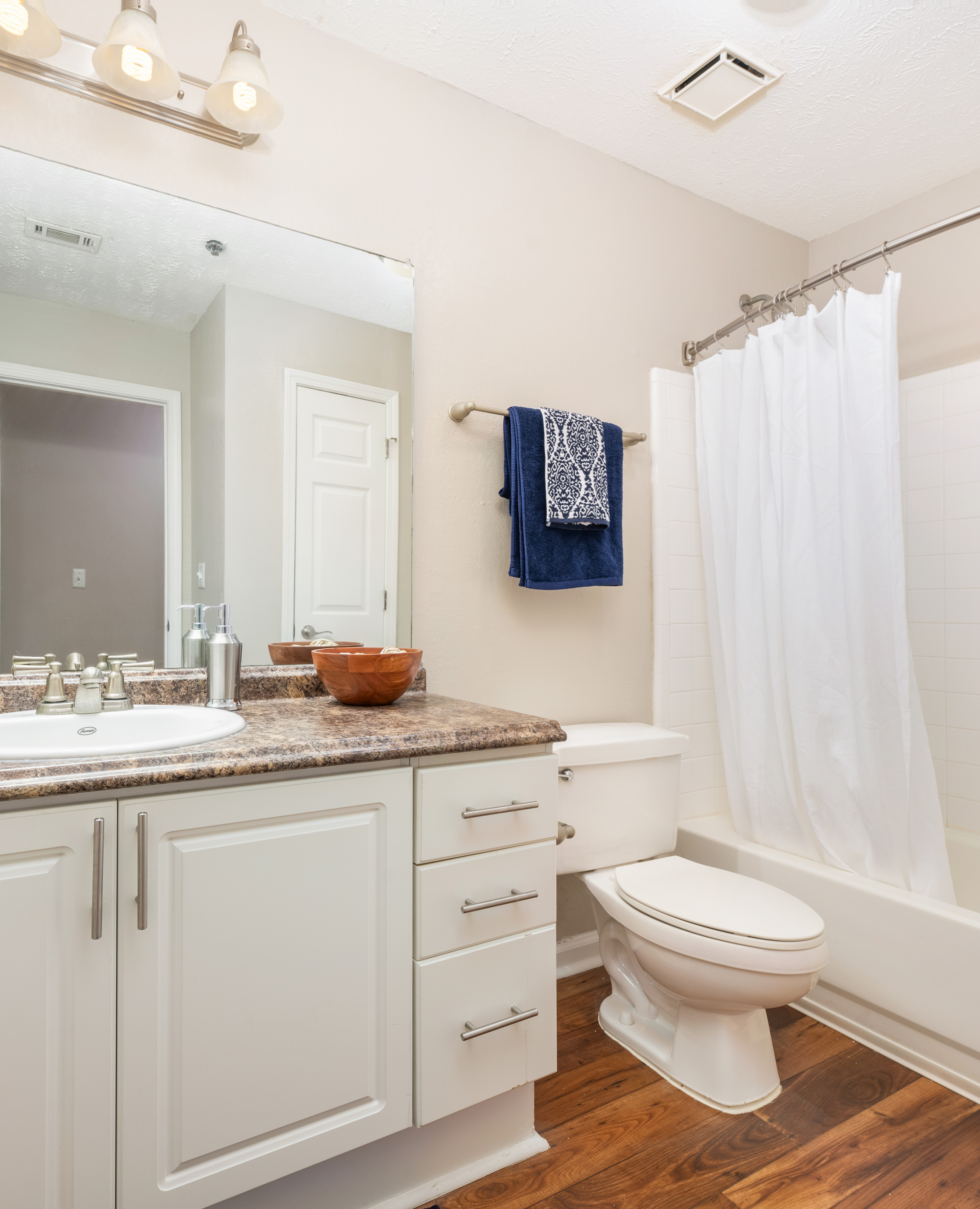 Bathroom with large vanity mirror with a single sink, dark marble countertops with white cabinets, and shower tub with curtains on wood floors at MAA Mount Vernon luxury apartments in Sandy Springs, GA