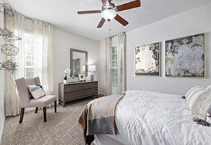 Bedroom with a ceiling fan, wall art and a white arm chair next to a dresser and windows at MAA Pleasant Hill luxury apartments in Duluth, GA