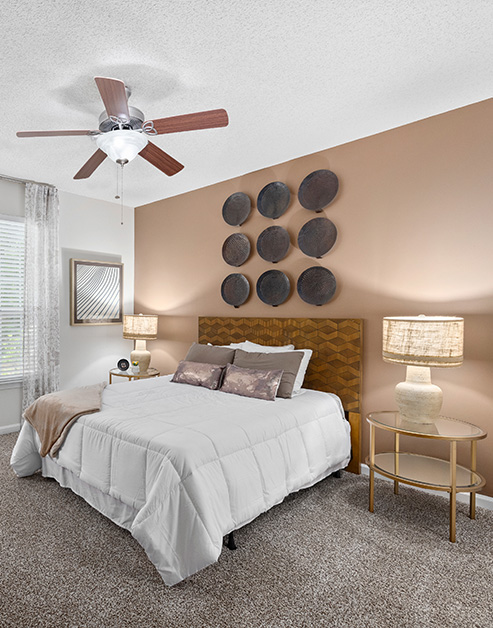 Model bedroom with a ceiling fan, a wood headboard, tan accent wall with circular wall art and a night stand at MAA Pleasant Hill luxury apartments in Duluth, GA