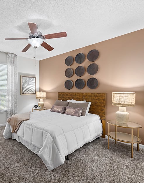 Model bedroom with a ceiling fan, a wood headboard, tan accent wall with circular wall art and a night stand at MAA Pleasant Hill luxury apartments in Duluth, GA