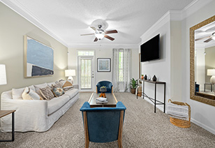 Side view of model living room with a ceiling fan, white couch and plush blue arm chair sitting area and a wall-mounted TV on grey carpeted floors at MAA Pleasant Hill luxury apartments in Duluth, GA