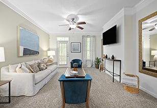 Side view of model living room with a ceiling fan, white couch and plush blue arm chair sitting area and a wall-mounted TV on grey carpeted floors at MAA Pleasant Hill luxury apartments in Duluth, GA