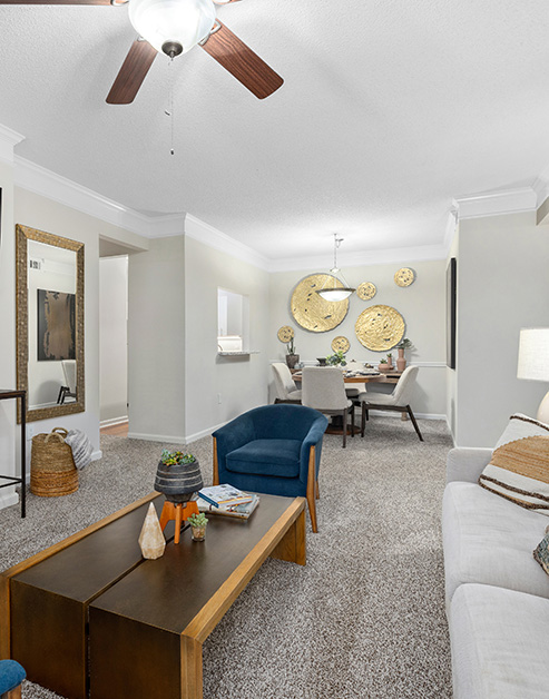 Model living room with a ceiling fan, gold circular wall art and wood rectangular coffee table with a plush blue armchair on grey carpeted floors at MAA Pleasant Hill luxury apartments in Duluth, GA