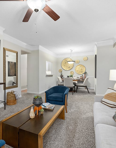Model living room with a ceiling fan, gold circular wall art and wood rectangular coffee table with a plush blue armchair on grey carpeted floors at MAA Pleasant Hill luxury apartments in Duluth, GA