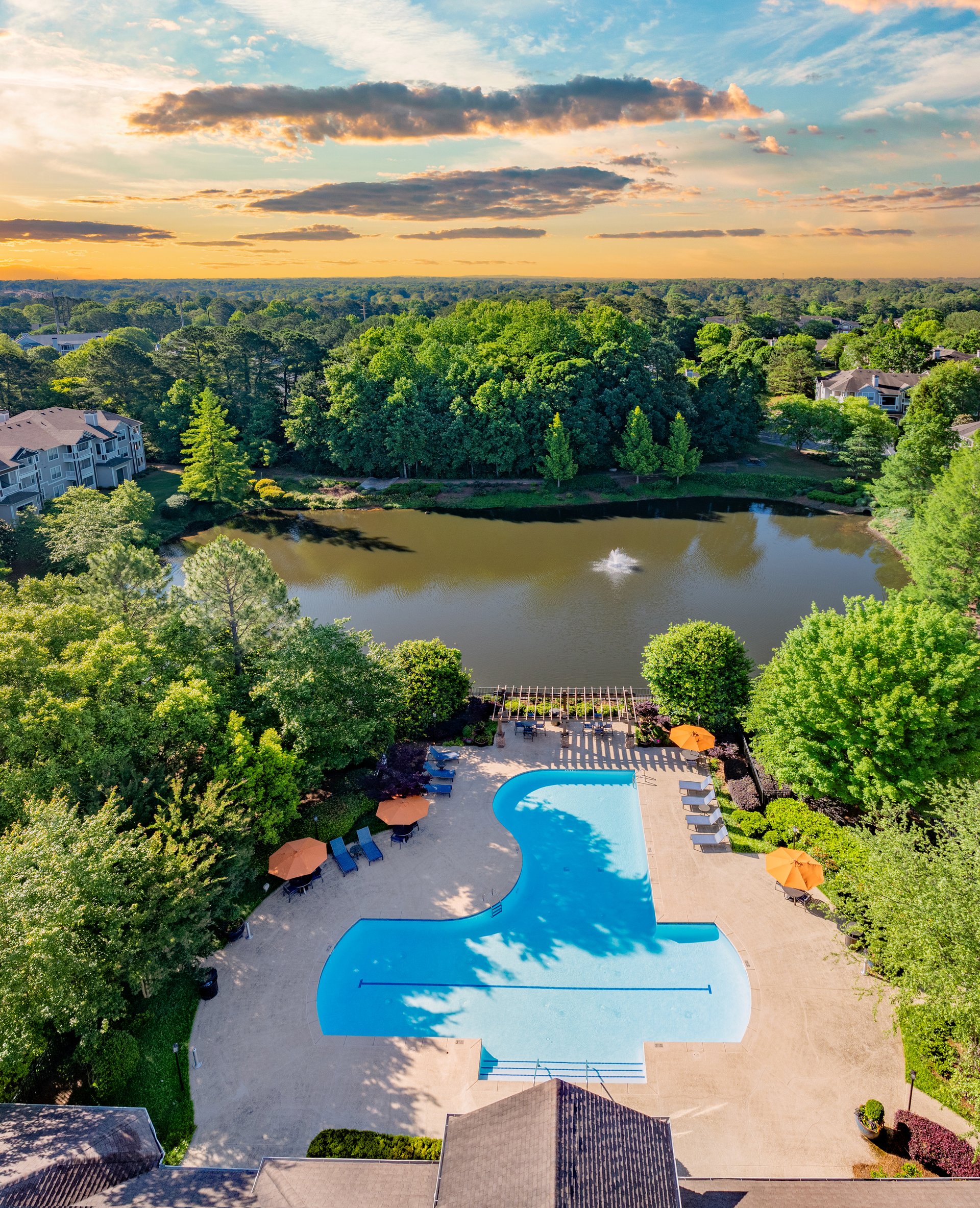 Aerial view of swimming pool with lounge chairs next to large lake surrounded by trees with a sunset sky at MAA Prescott luxury apartments in Duluth, GA