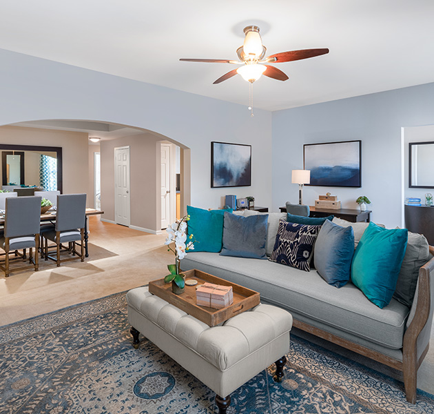 Model living room with arch entryway to dining area, with a ceiling fan and grey couch with accent pillows and ottoman on an area rug at MAA Riverside luxury apartments in Atlanta, GA