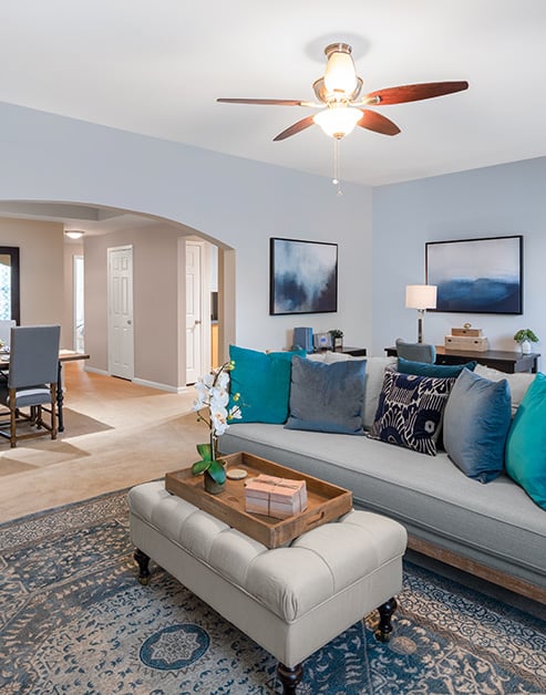 Model living room with arch entryway, a ceiling fan and grey couch with accent pillows next to an off-white ottoman on an area rug at MAA Riverside luxury apartments in Atlanta, GA