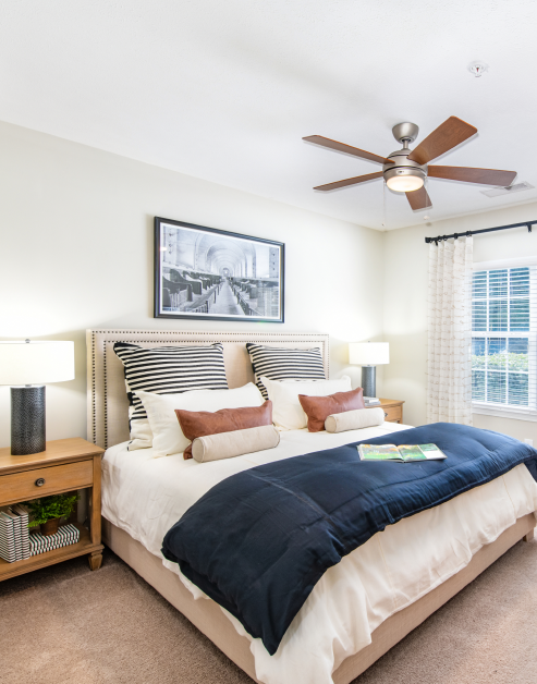 Bedroom with ceiling fan, wall art and night stands next to large window with blinds and curtains at MAA Ballantyne luxury apartments in Charlotte, NC