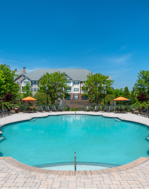 Long view of resort-style swimming pool with entrance steps and metal handrails, lounge chairs, sun umbrella with table and chairs at MAA Ballantyne luxury apartments in Charlotte, NC