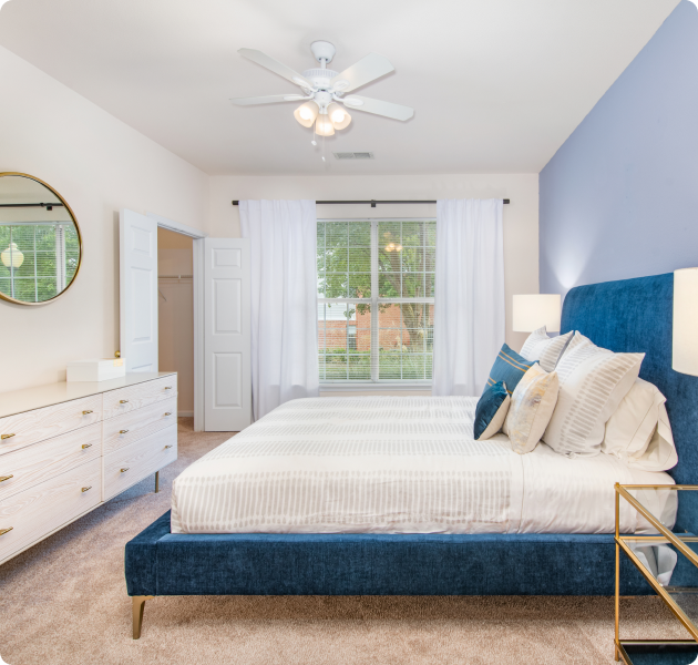 An apartment model bedroom with ceiling fan, round mirror above white dresser next to plush blue-framed bed and a large window with curtains at MAA Beverly Crest luxury apartments in Charlotte, NC
