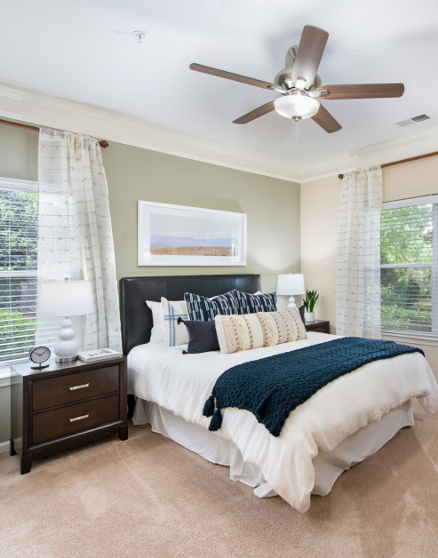 Model bedroom with ceiling fan, queen box bed with black headboard and wooden night stand next to windows with blinds at MAA Cornelius luxury apartments in Cornelius, NC