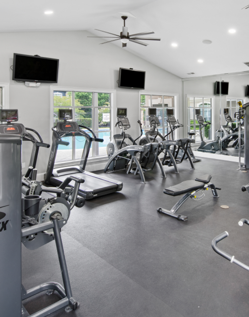 Fitness center with treadmills in natural light next to large windows, wall mounted TVs and ceiling fan with vaulted ceiling at MAA Cornelius luxury apartments in Cornelius, NC