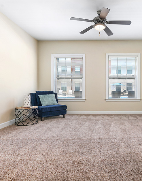 Empty carpeted bedroom with a dark blue armchair and side table, ceiling fan and windows with blinds at MAA Enclave luxury apartments in Charlotte, NC