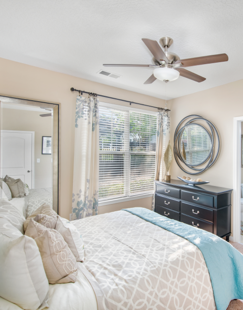 Bedroom with ceiling fan, large window with blinds and curtains next to a wall-mounted round mirror at MAA Huntersville luxury apartments in Huntersville, NC