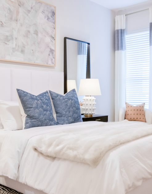 Bedroom with wall painting above the white queen bed, a bedside table lamp under mirror next to a window at MAA LoSo luxury apartments in Charlotte, NC