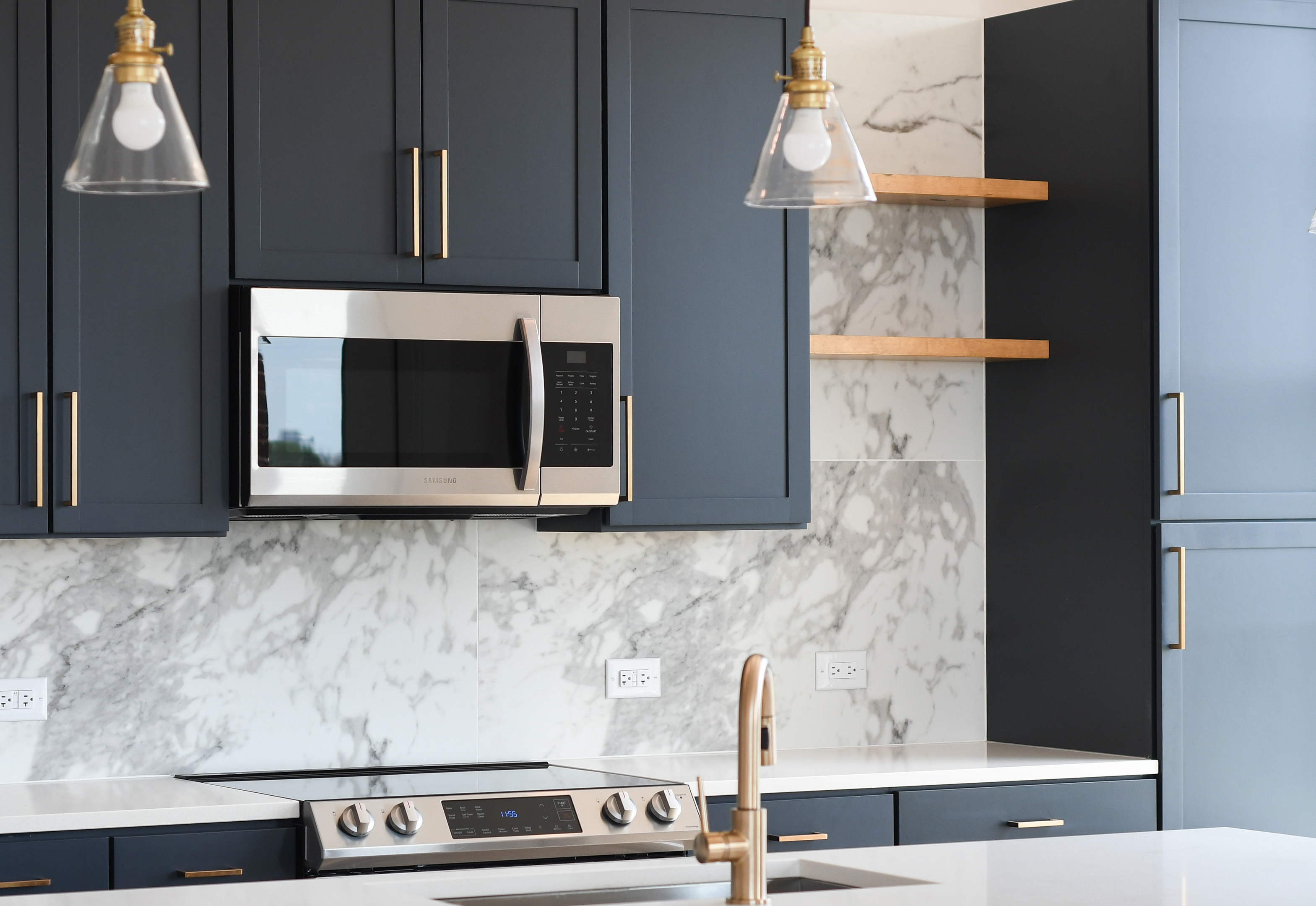 Close up of kitchen marble backsplash wall with dark gray cabinets and floating shelving, stainless-steel appliances and hanging light fixtures over countertop at MAA LoSo luxury apartments in Charlotte, NC