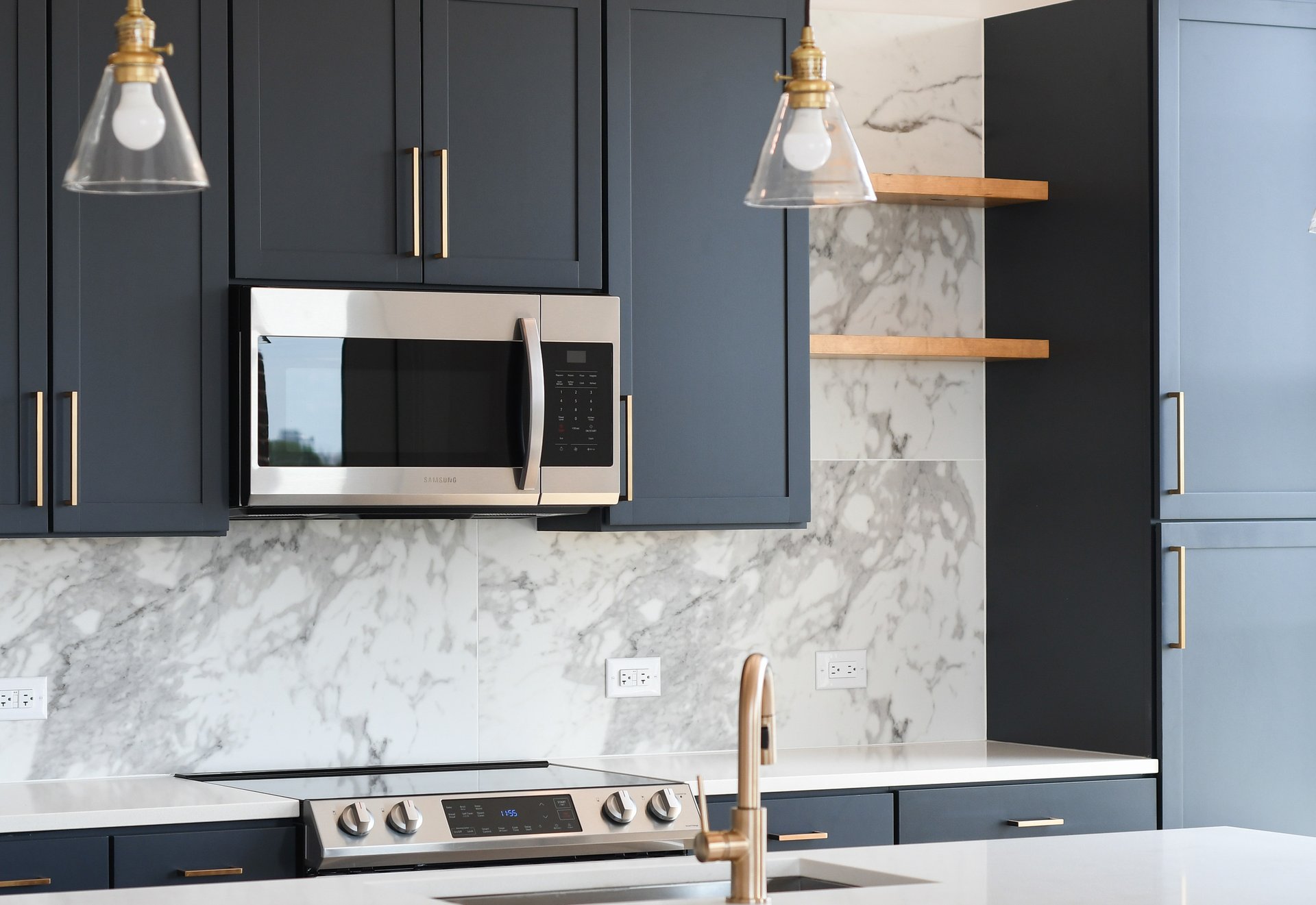 Close up of kitchen marble backsplash wall with dark gray cabinets and floating shelving, stainless-steel appliances and hanging light fixtures over countertop at MAA LoSo luxury apartments in Charlotte, NC
