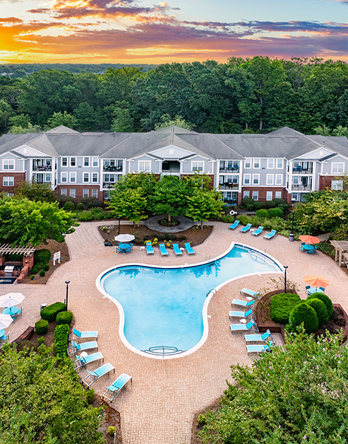 Aerial view of resort-style pool with lounge chairs and lush greenery situated next to the apartment building at MAA Matthews Commons luxury apartments in Matthews, NC