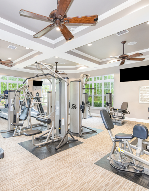 Fitness center with ceiling fans, wall mounted TVs and various workout machines in natural light next to large windows at MAA Matthews Commons luxury apartments in Matthews, NC
